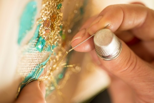 Close up shot of a ladies hands wearing a thimble and doing embroidery with gold thread and sequins