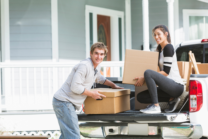 A young couple moving into a house or apartment. Their pickup truck is full of cardboard boxes and furniture. They are smiling at the camera, lifting the boxes.