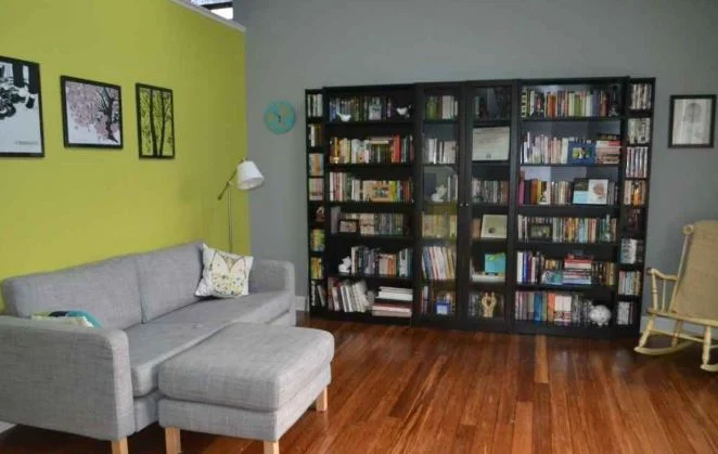 A room with bookshelf, grey couch, and warm wood flooring